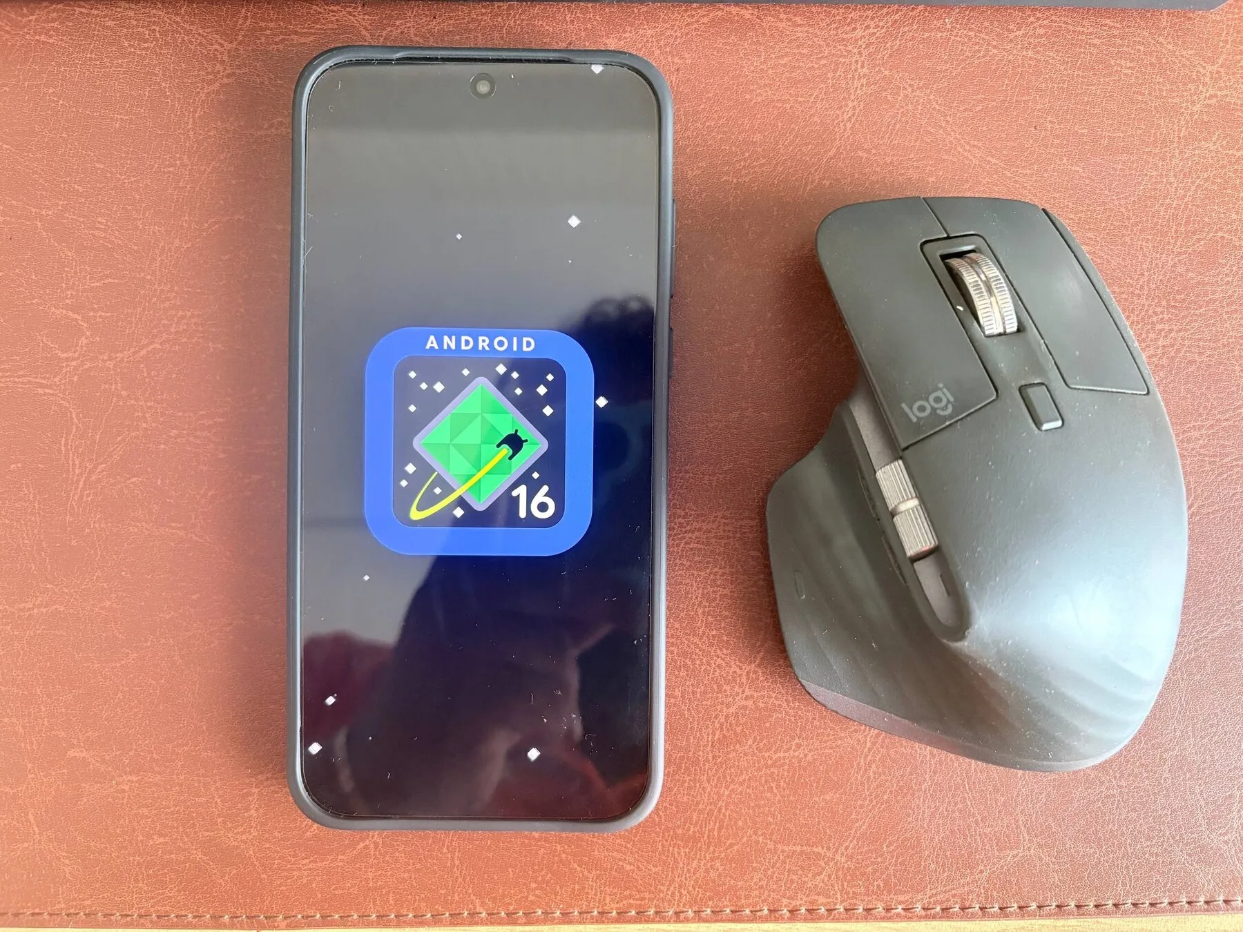 A OnePlus phone displaying the Android 16 easter egg beside an MX Master mouse
